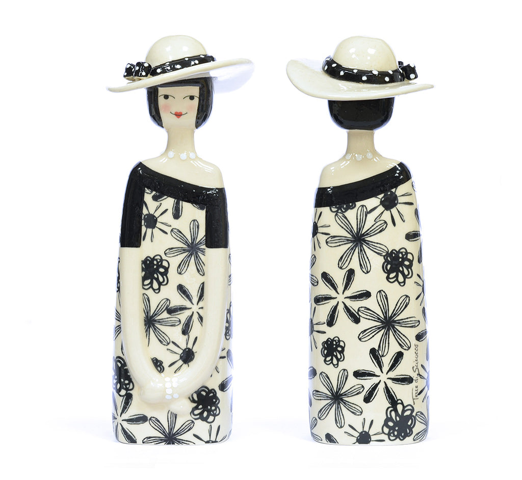Two ceramic figurines of women shown from the front and back, wearing wide-brimmed white hats with black patterns and long black dresses with a black floral print on an off-white background.