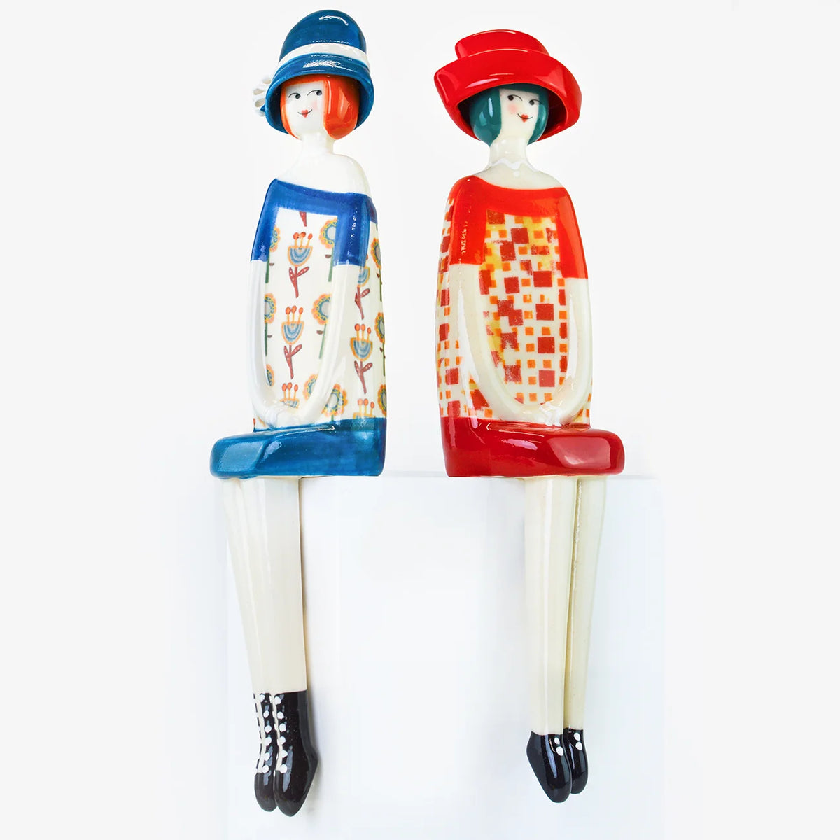 Two decorative seated figurines of women in colorful retro-style clothing, one in blue and one in red, displayed against a plain white background.