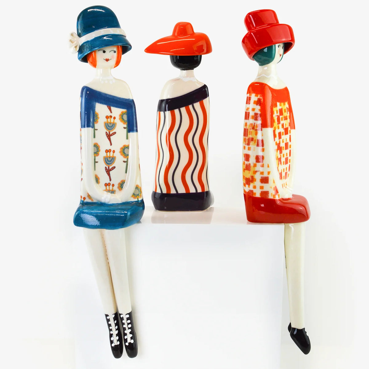 Three stylized seated figurines dressed in colorful retro outfits, including hats, dresses, and patterned clothing, arranged side by side on a white surface against a white background.