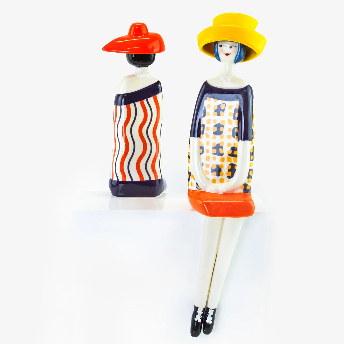 Two stylized ceramic figurines of women in colorful patterned dresses and hats, one shown from the back and one from the front, on a white background.