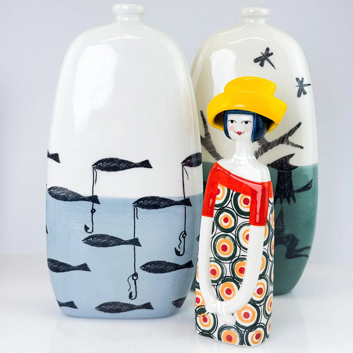 Two decorative ceramic vases with a stylized figurine in front. The vases are white with black fish and fishing line motifs on a light blue lower half, while the figurine wears a bright yellow hat, a red top, and a black, white, orange, and red patterned skirt.