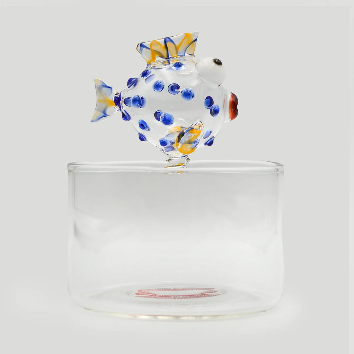 A hand-blown glass fish sculpture with blue, yellow, and white patterns, perched on top of a clear glass small bowl.