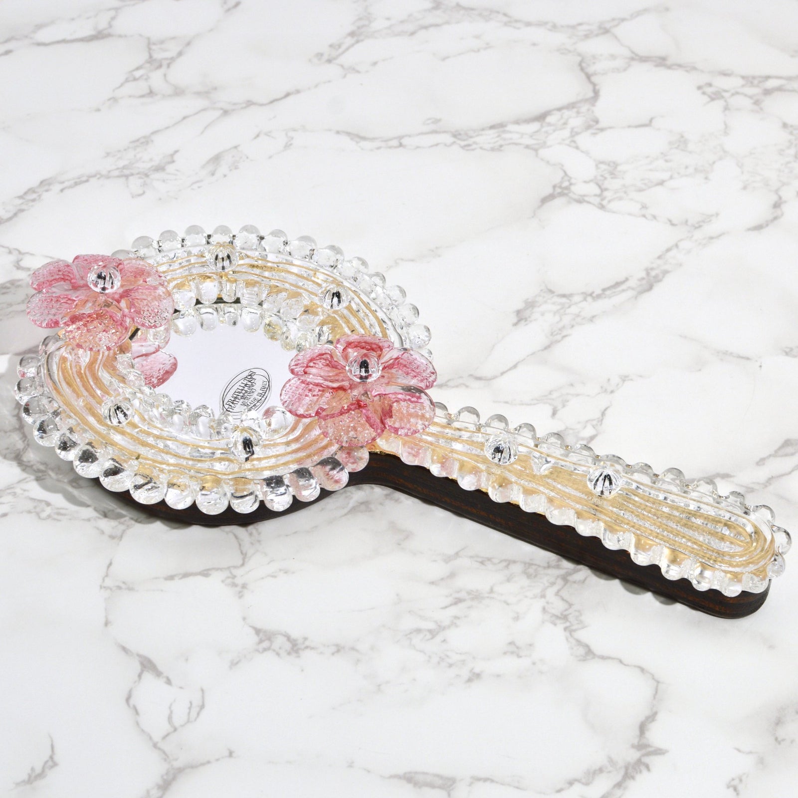 Murano glass hand mirror with pink or clear flower designs, made in Italy, featuring a decorative floral pattern and a reflective surface.