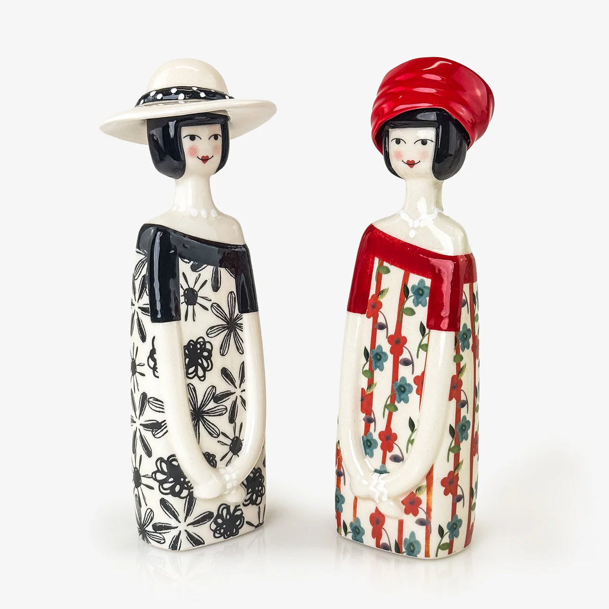 Two decorative ceramic figurines of women standing side by side on a white background. The figurine on the left wears a white hat with black trim and a black-and-white patterned dress.