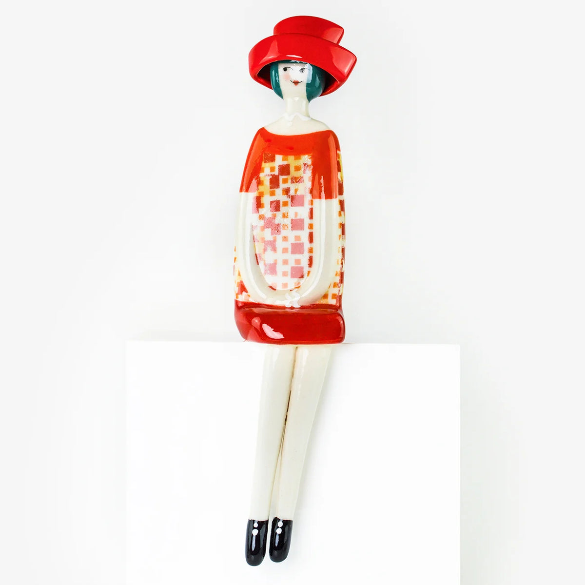 A stylized ceramic figurine of a seated woman wearing a red hat and a red-and-white patterned dress, displayed against a clean white background.