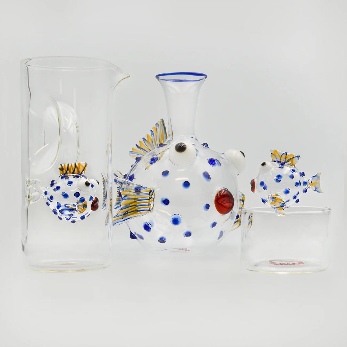A set of hand-blown glass pufferfish-shaped glass carafe with blue dots, yellow-striped fins, and a red glass lips accompanied by a tall matching glass pitcher, a small bowl, with a fish perched on it's ledge.