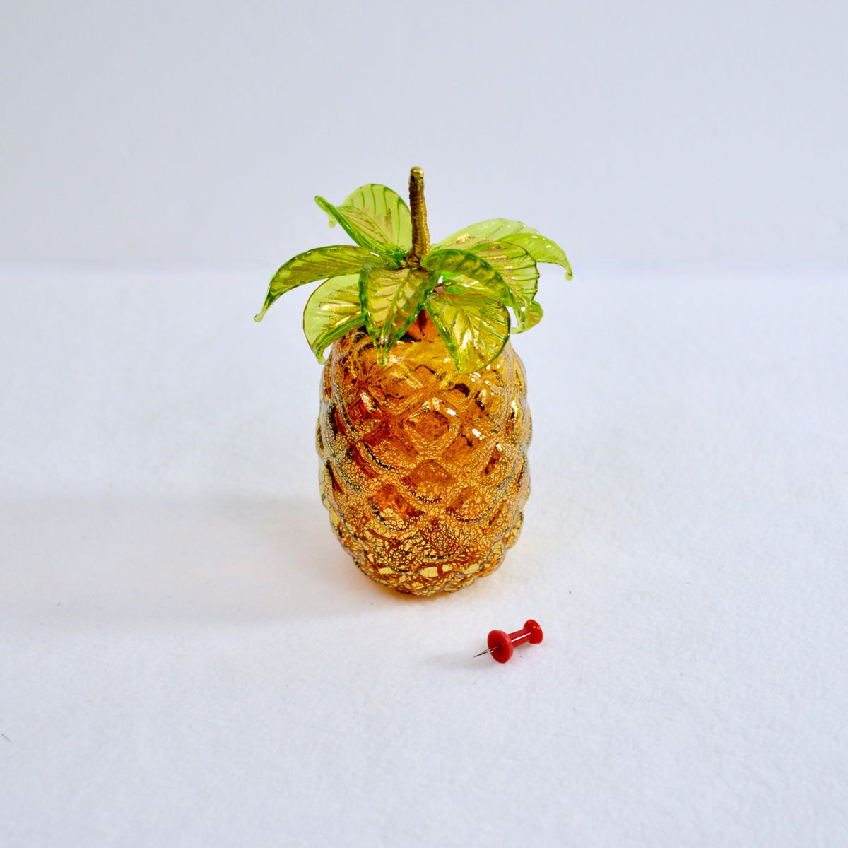 An amber colored Murano glass pineapple is shown with a push pin for scale.