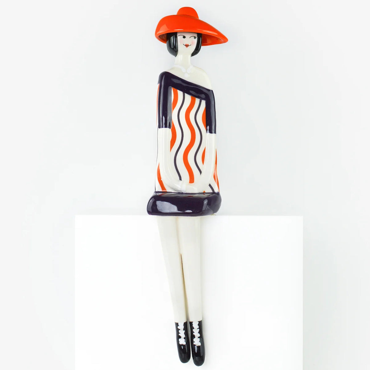 A stylized ceramic figurine of a seated woman with a wide-brimmed orange hat, a white dress with orange and black wavy stripes, and black-and-white shoes, displayed on a white pedestal against a white background.