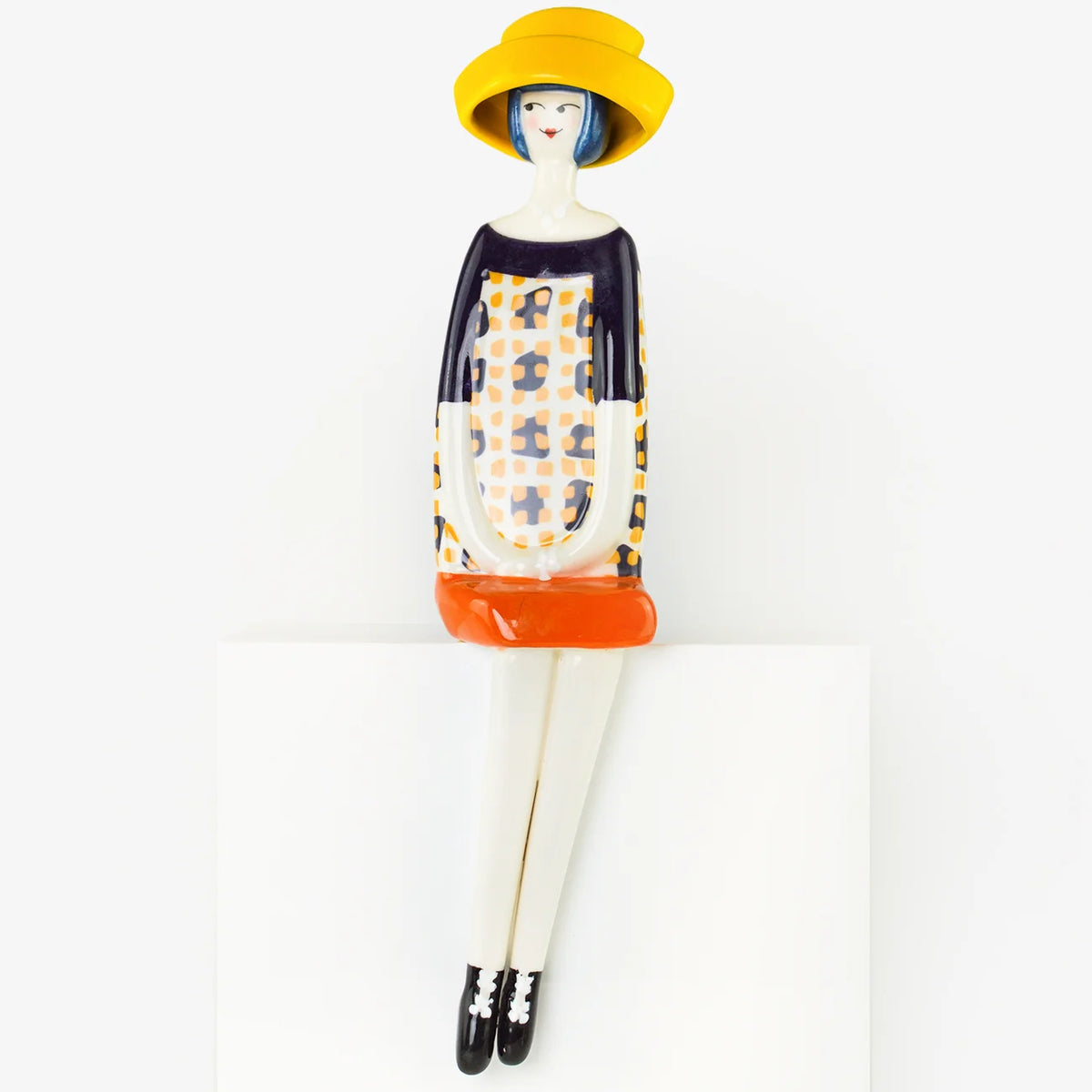 A stylized ceramic figurine of a seated woman wearing a wide-brimmed yellow hat, a patterned dress with orange and navy sections, and black-and-white shoes, displayed against a clean white background.