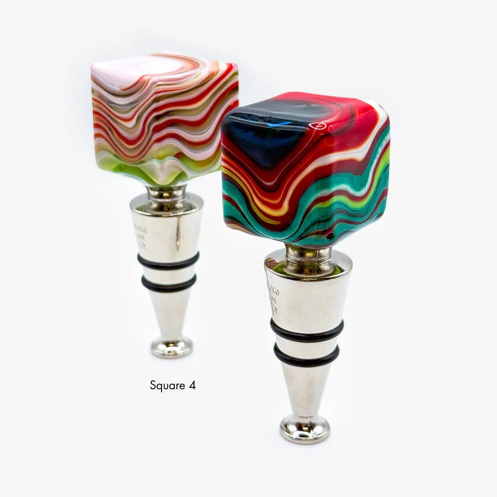 Two square-shaped wine bottle stoppers with colorful marbled swirls in red, green, blue, yellow, and white, set on silver metal bases against a light gray background.