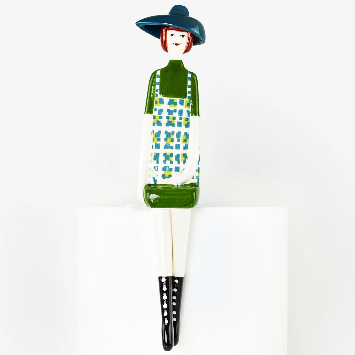 A stylized ceramic figurine of a woman wearing a dark blue hat, green top, white apron with blue and green check pattern, and black boots, seated on a white block against a white background.