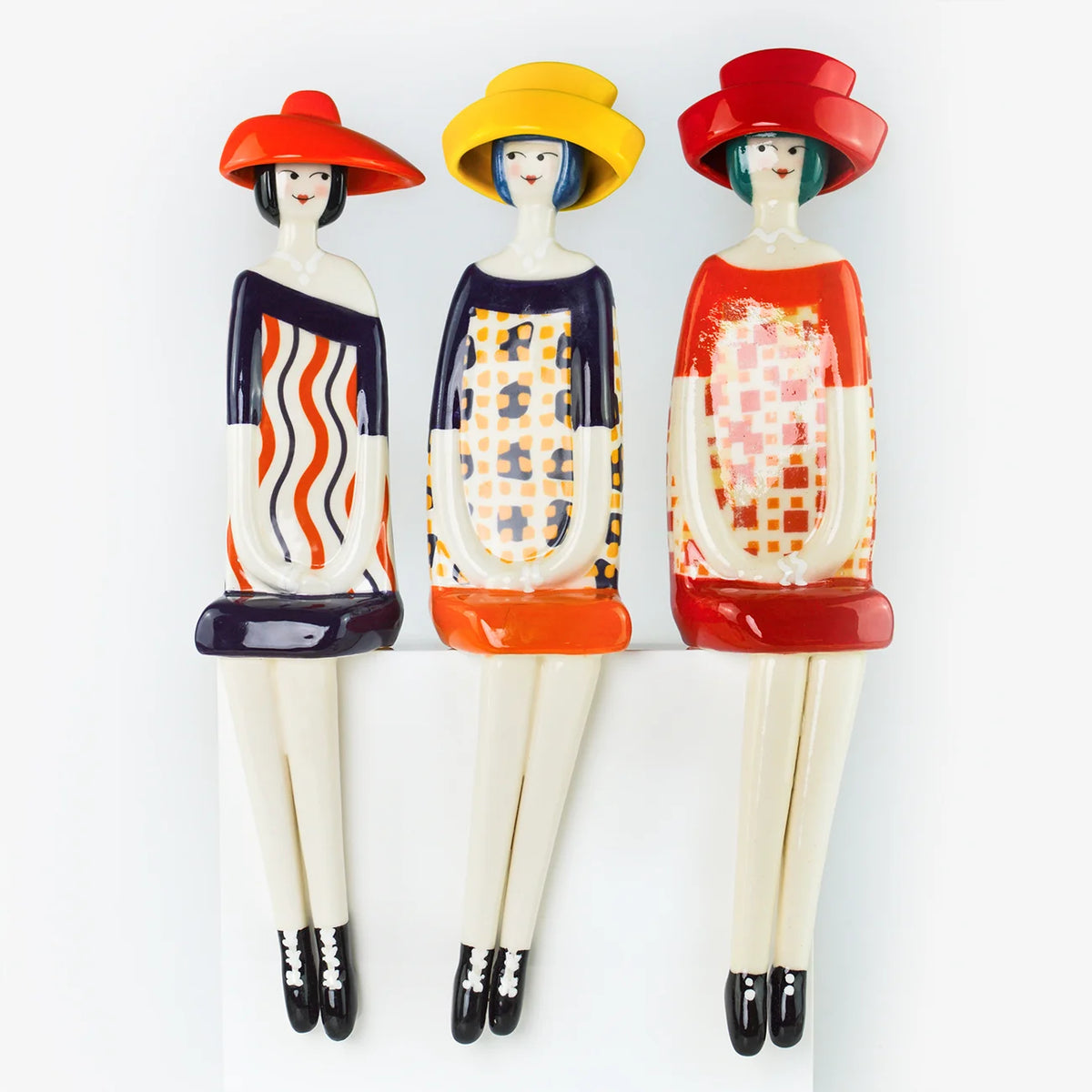 Three decorative figurines of women in retro 1960s style, wearing colorful hats and patterned dresses, mounted on a white wall.