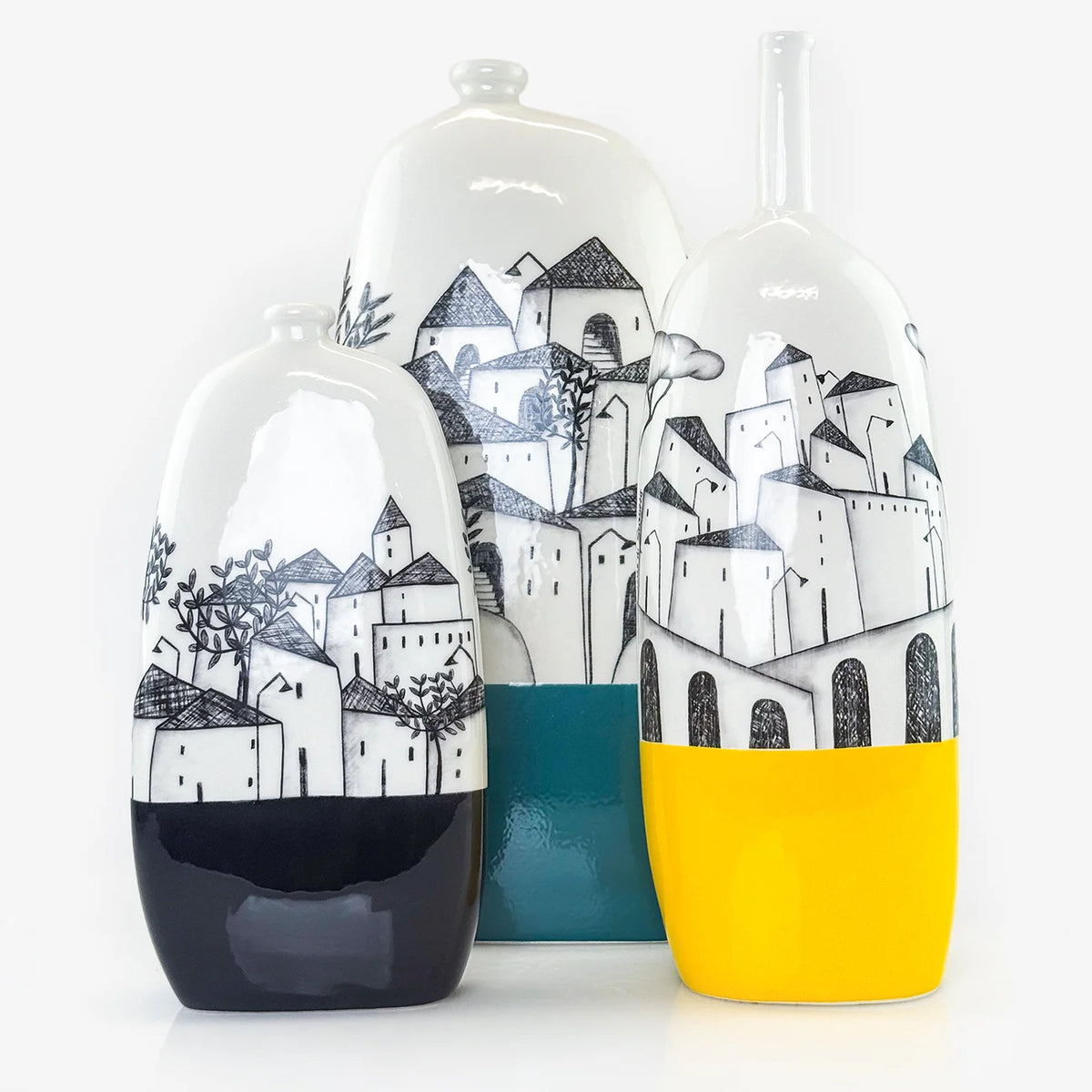 Three ceramic decorative vases with hand-painted black and white village scenes, displayed on a clean white background. The left vase has a black base, the center vase has a teal base, and the right vase has a bright yellow base.