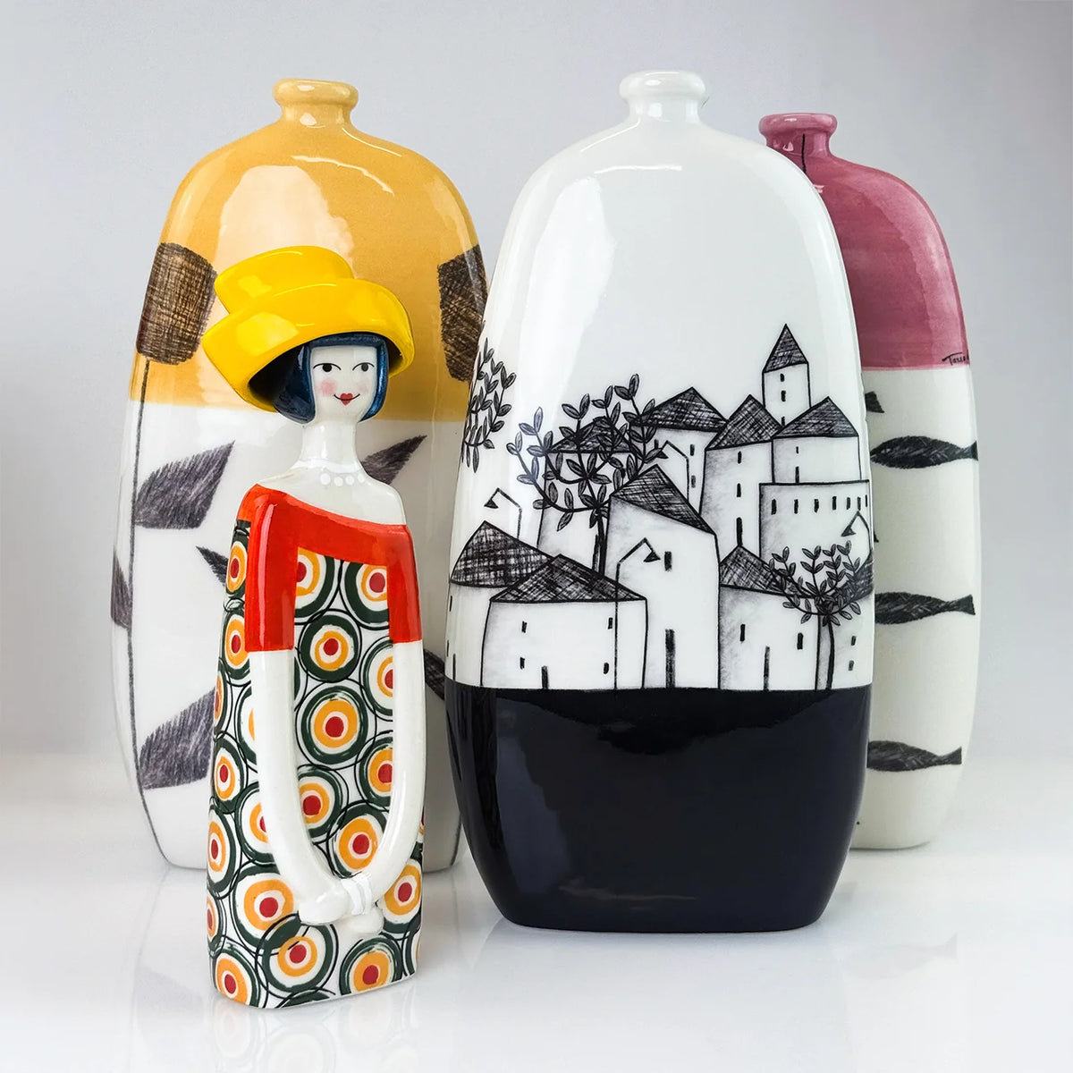 Three decorative ceramic vases with a stylized figurine in the foreground, photographed on a clean light gray studio background. The vases have bold abstract and landscape-style painted patterns in white, black, yellow, pink, red, orange, and gray.