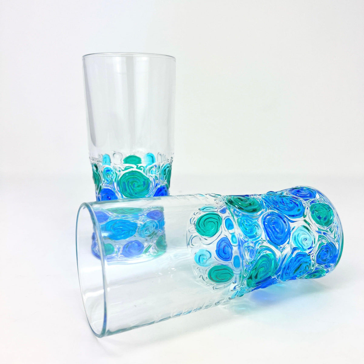 Two Italian Crystal tall drinking glasses with bright blue, green, and turquoise swirls of color on the bottom half. One glass lays on it&#39;s side, the other stands upright.